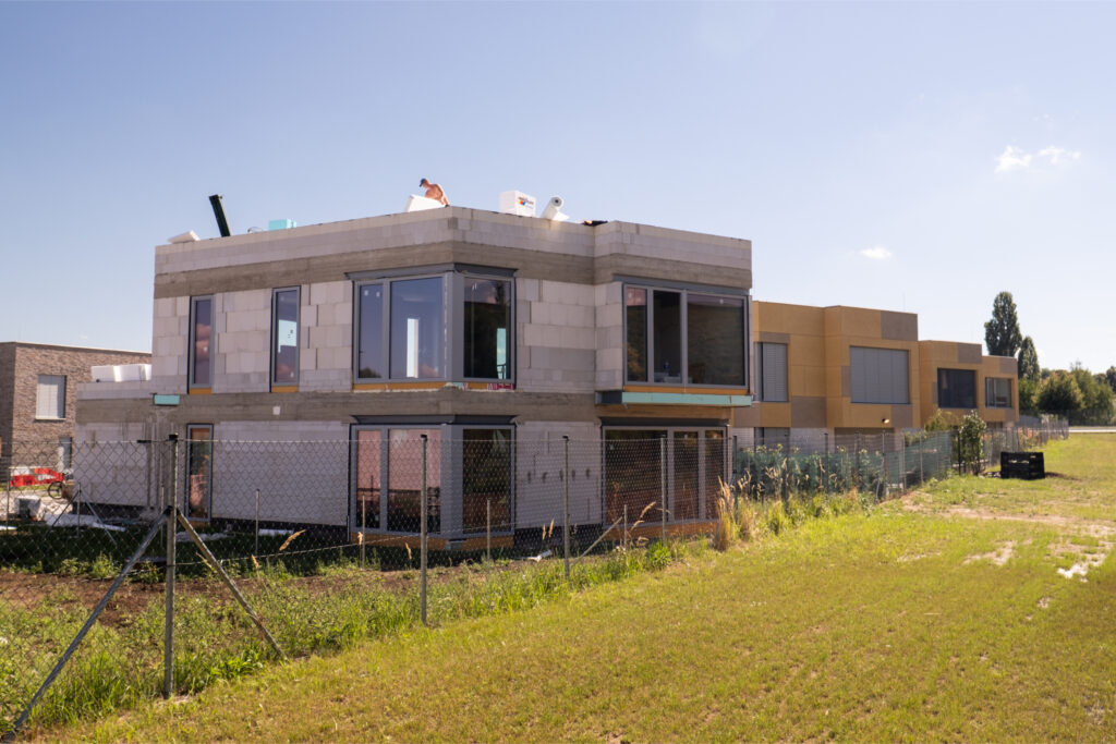 Hrubá stavba dvoupodlažního rodinného domu v novém developerském projektu, bez fasády a finálních úprav, obklopená nezakultivovaným pozemkem.