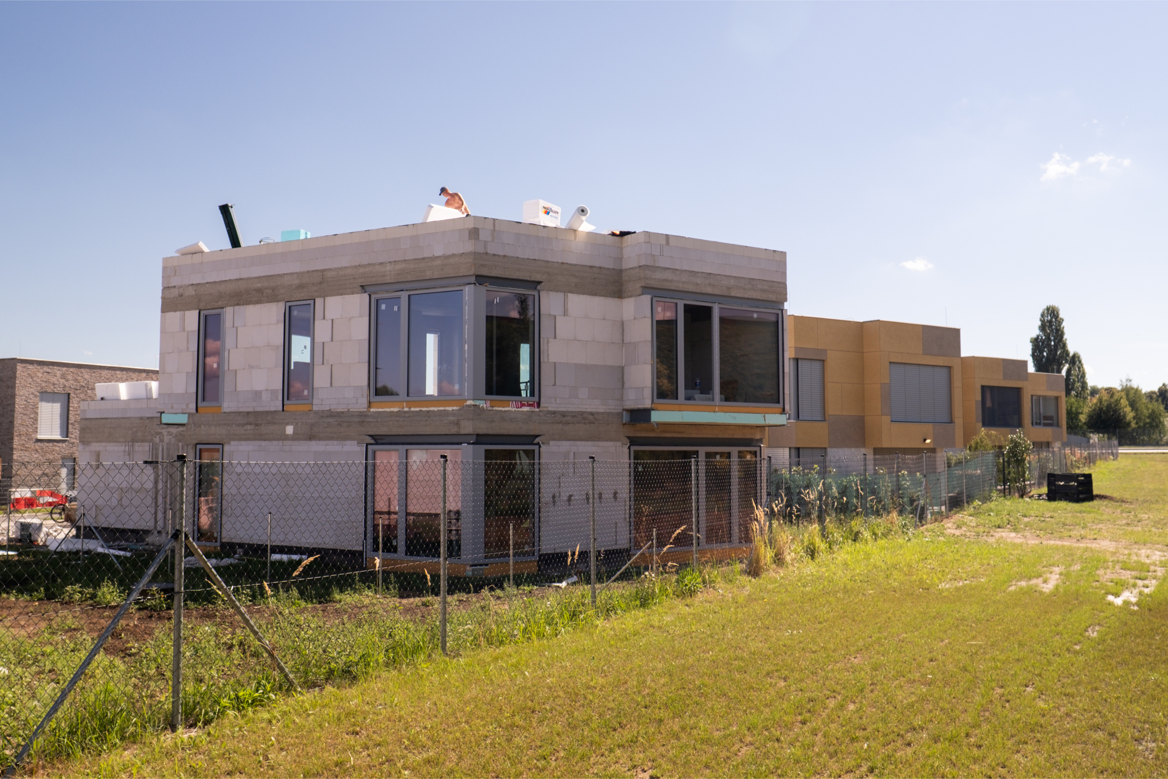 Hrubá stavba dvoupodlažního rodinného domu v novém developerském projektu, bez fasády a finálních úprav, obklopená nezakultivovaným pozemkem.