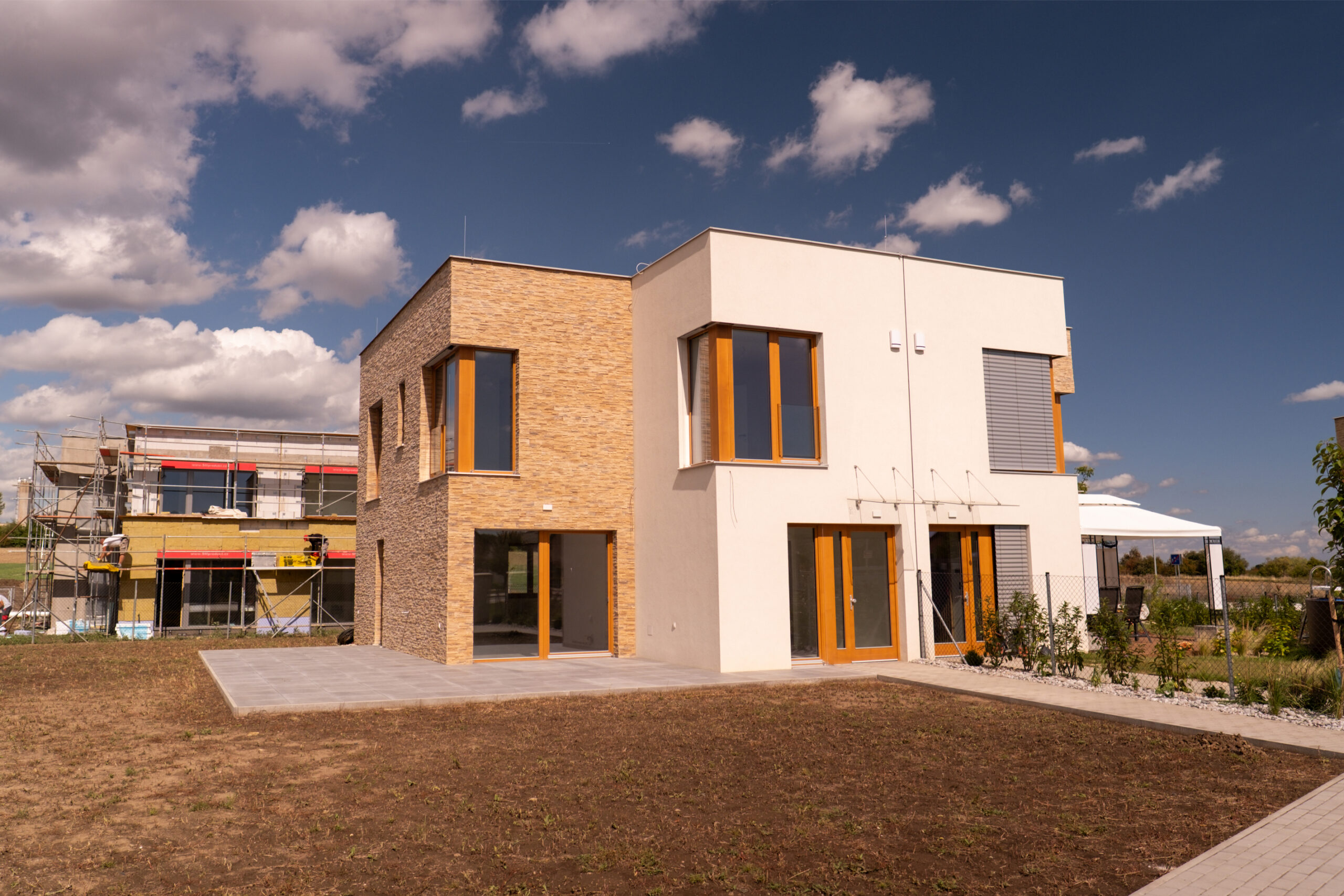 Moderní nízkoenergetický rodinný dům s plochou střechou, kombinací bílé a dřevěné fasády, velkými okny a venkovním stíněním; novostavba v rezidenční lokalitě za slunného dne. Fotografie ukazuje charakter kvalitně postavené novostavby – čisté detaily fasády, velkoformátová okna pro dostatek světla, promyšlené venkovní stínění a terasu jako propojení interiéru se zahradou; příklad developerského projektu v Praze a okolí.