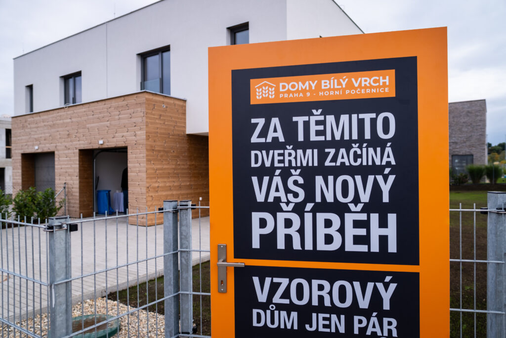 Vstup k projektu Domy Bílý Vrch s cedulí „Za těmito dveřmi začíná váš nový příběh“ u vzorového domu v Praze – Horních Počernicích.